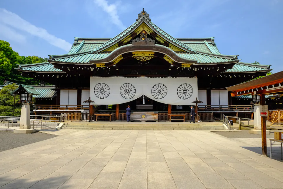 A traditional Japanese shrine with a green-tiled roof, intricate woodwork, and large white banners displaying circular patterns. The shrine entrance is framed by a spacious, paved courtyard, with a few visitors standing near the entrance. Blue sky in the backdrop.