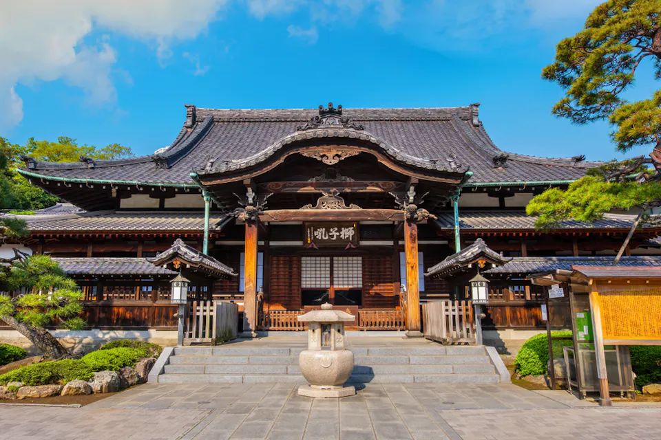 A traditional Japanese temple with intricate wooden architecture and a tiled roof. A stone lantern stands at the front, and two trees frame the entrance. Steps lead up to the main structure, and clear skies with white clouds are visible in the background.