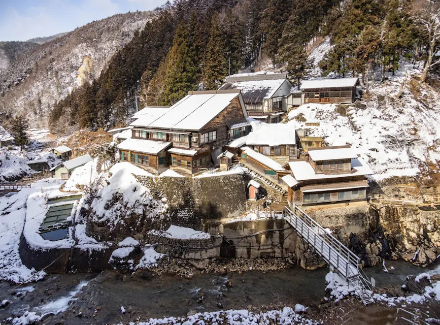 A scenic view of snow-covered traditional wooden houses nestled on a hillside, surrounded by a dense forest. A river flows in the foreground, with a small bridge crossing it, connecting to the houses. Snow blankets the roofs and the ground, creating a serene winter atmosphere.