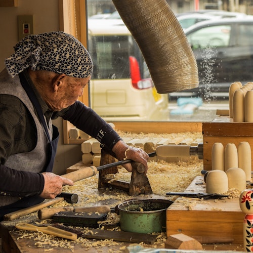 Hands-On Workshops for Artisanal Souvenirs in Japan An elderly person wearing a blue bandana works in a woodshop, carving wooden objects with a chisel and mallet. The workbench is covered in wood shavings, and several finished wooden dolls are displayed in the foreground. A car is visible through the window.