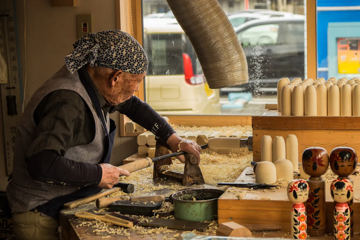 Hands-On Workshops for Artisanal Souvenirs in Japan An elderly person wearing a blue bandana works in a woodshop, carving wooden objects with a chisel and mallet. The workbench is covered in wood shavings, and several finished wooden dolls are displayed in the foreground. A car is visible through the window.