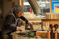 An elderly person wearing a blue bandana works in a woodshop, carving wooden objects with a chisel and mallet. The workbench is covered in wood shavings, and several finished wooden dolls are displayed in the foreground. A car is visible through the window.