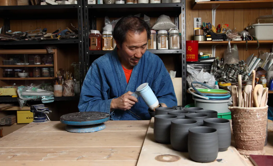 A man wearing a blue robe is seated at a wooden table, working on a light blue ceramic piece in a pottery studio. Several black clay pots and various tools are spread across the table, with shelves holding additional pottery materials and supplies in the background.