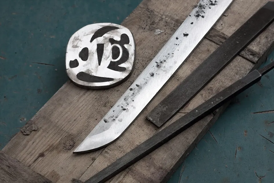 A partially polished knife blade, an oval metal plate with markings, and two metal files lie on a wooden plank against a blue background, suggesting a scene of knife crafting or sharpening.