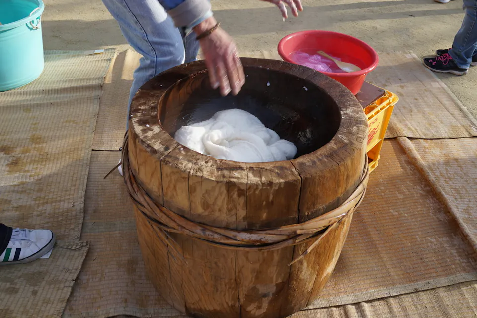 A person kneads freshly pounded rice dough in a large wooden mortar (usu) typically used for mochi making. A red bowl with additional dough and water is nearby, set on a crate, while a towel and other items lie around the setup.
