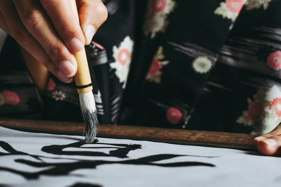 A person wearing a floral-patterned garment uses a brush to write traditional calligraphy on a piece of paper. The close-up shot focuses on the hand holding the brush and the partially completed black ink characters on the paper.