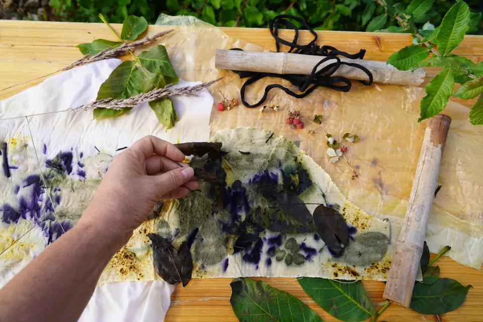 A person arranges leaves, flowers, and plant materials onto a piece of fabric laid out on a wooden table. The fabric displays imprints of purple and green colors from the plants. Nearby are various leaves, seeds, wooden sticks, and black strings.