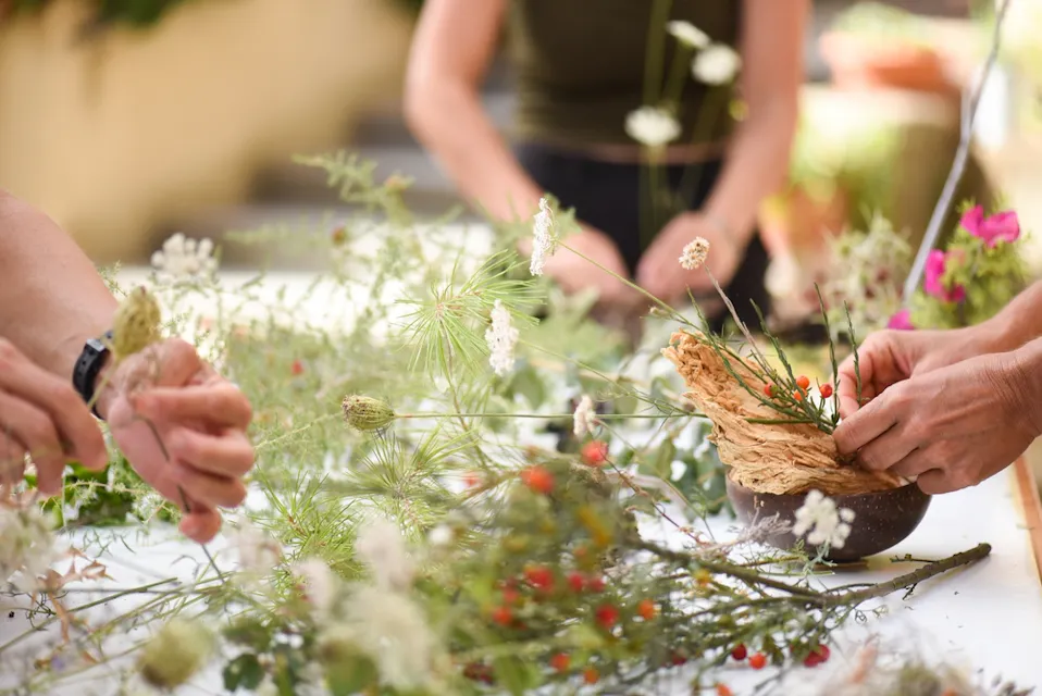 Three people are arranging various flowers and greenery on a table. Their hands are visible as they work with delicate white blooms, green stems, and some red berries. One person is holding a beige, textured material, possibly dried leaves or petals.