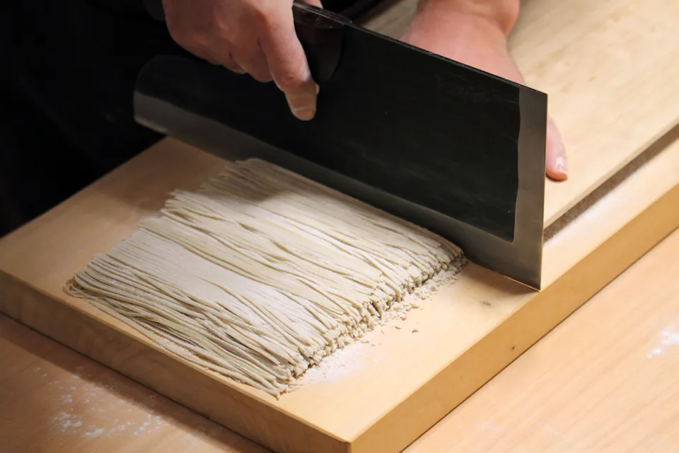 A person slices thin, uniform strips of raw noodle dough with a large, rectangular knife on a wooden surface dusted with flour.