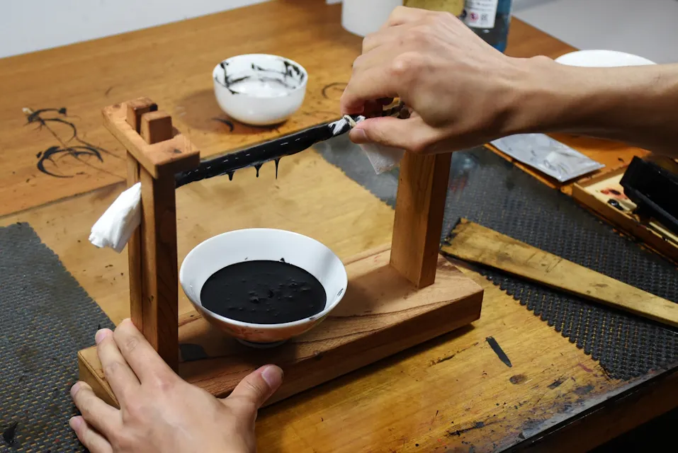 A person grinds ink using an ink stone and a wooden frame support. A bowl filled with black ink is placed beneath the setup on a wooden table. The person is holding the ink stick and pressing it against the stone, which is mounted on the wooden frame.