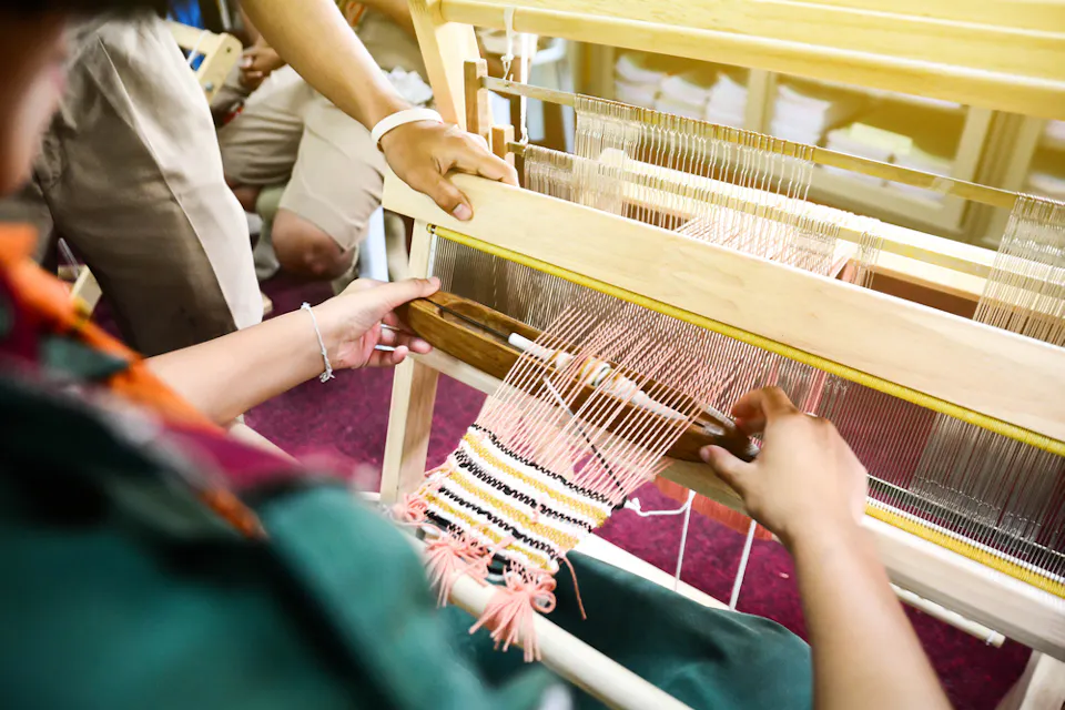 A person is weaving fabric using a traditional wooden loom. Their hands are guiding the shuttle through the threads, with partially woven, colorful fabric visible. Other people, blurred in the background, are observing the process.