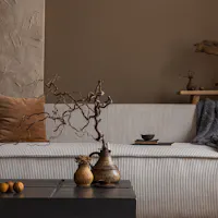 Wabi-Sabi A cozy, minimalistic living room scene with a textured beige sofa adorned with brown and beige cushions and a gray throw blanket. A rustic wooden shelf with decorative items is in the background. A black coffee table with ornamental vases and nuts is in the foreground.