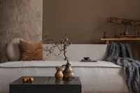 A cozy, minimalistic living room scene with a textured beige sofa adorned with brown and beige cushions and a gray throw blanket. A rustic wooden shelf with decorative items is in the background. A black coffee table with ornamental vases and nuts is in the foreground.