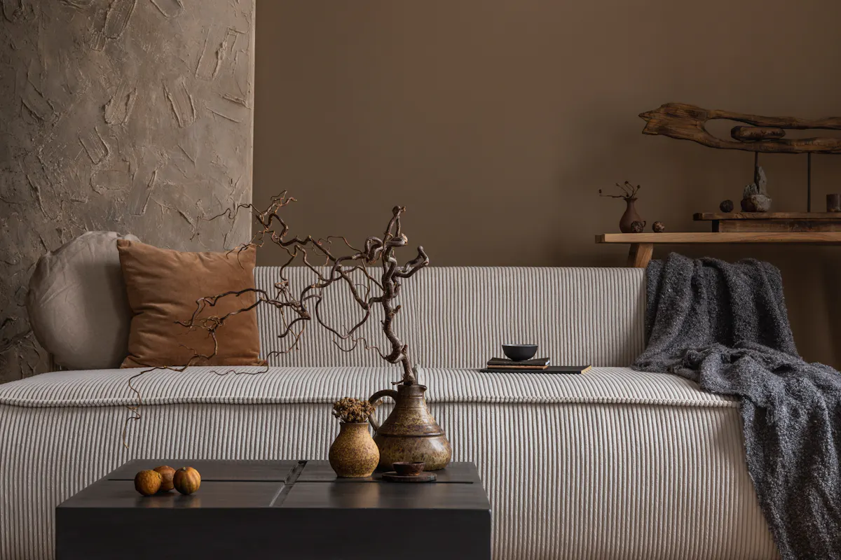 A cozy, minimalistic living room scene with a textured beige sofa adorned with brown and beige cushions and a gray throw blanket. A rustic wooden shelf with decorative items is in the background. A black coffee table with ornamental vases and nuts is in the foreground.