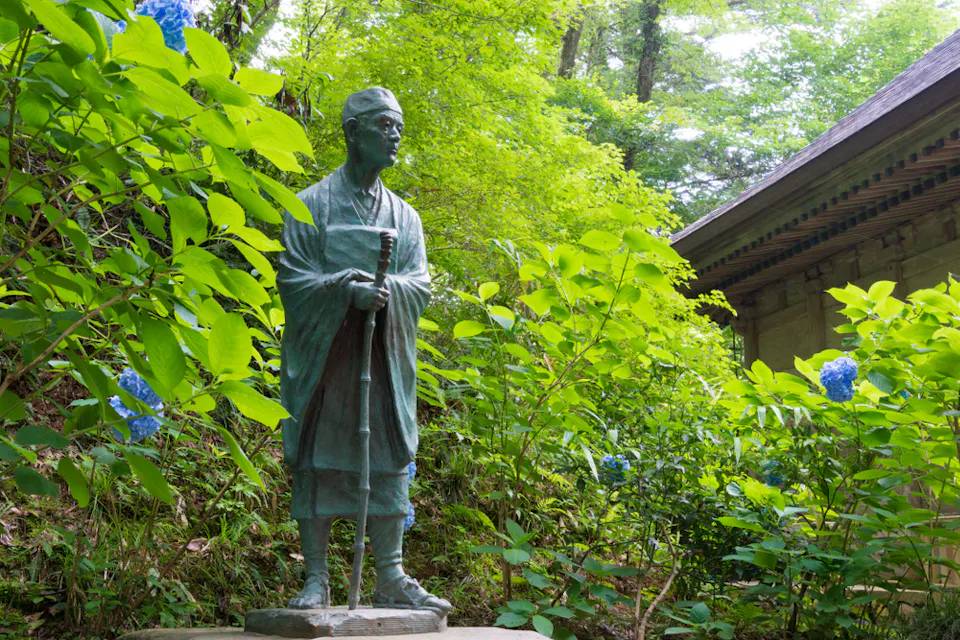 Matsuo Basho A bronze statue of a person wearing traditional clothing stands amid lush green foliage and blooming blue hydrangeas. The statue is positioned beside a wooden building with ornate eaves, all set in a serene, forested environment.
