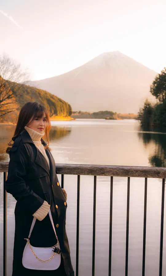 Lake Tanuki A woman in a black coat stands by a railing overlooking a calm lake, with Mount Fuji in the background, surrounded by trees and bathed in warm, soft light.