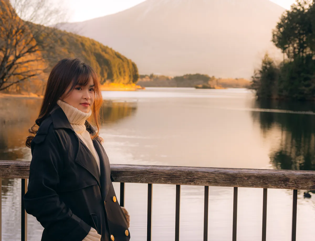 Lake Tanuki A woman in a black coat stands by a railing overlooking a calm lake, with Mount Fuji in the background, surrounded by trees and bathed in warm, soft light.