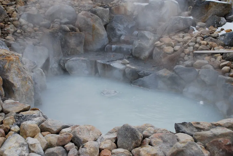 A small, steaming hot spring pool surrounded by an assortment of rocks. The water is light blue due to minerals, and steam is rising, indicating its warmth. In the background, there is a gentle stone step formation leading into the pool.
