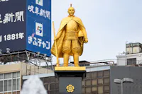 A golden statue of a historical samurai warrior stands on a black pedestal adorned with a floral emblem. The statue is dressed in traditional armor and holds a sword. Behind the statue, there are modern buildings and billboards with Japanese text.
