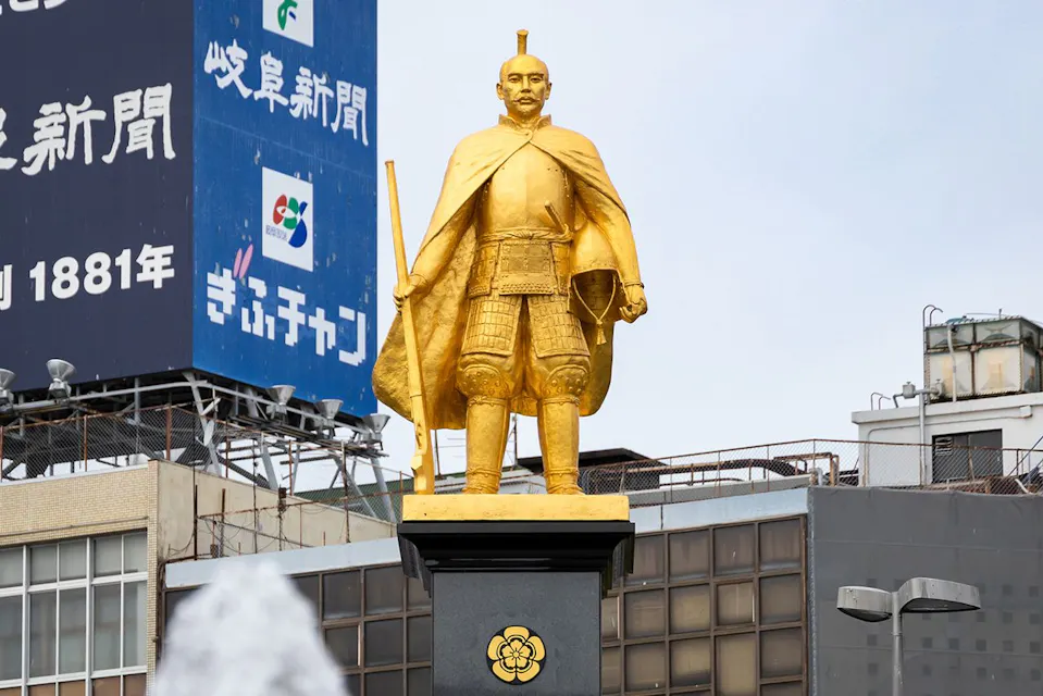 A golden statue of a historical samurai warrior stands on a black pedestal adorned with a floral emblem. The statue is dressed in traditional armor and holds a sword. Behind the statue, there are modern buildings and billboards with Japanese text.