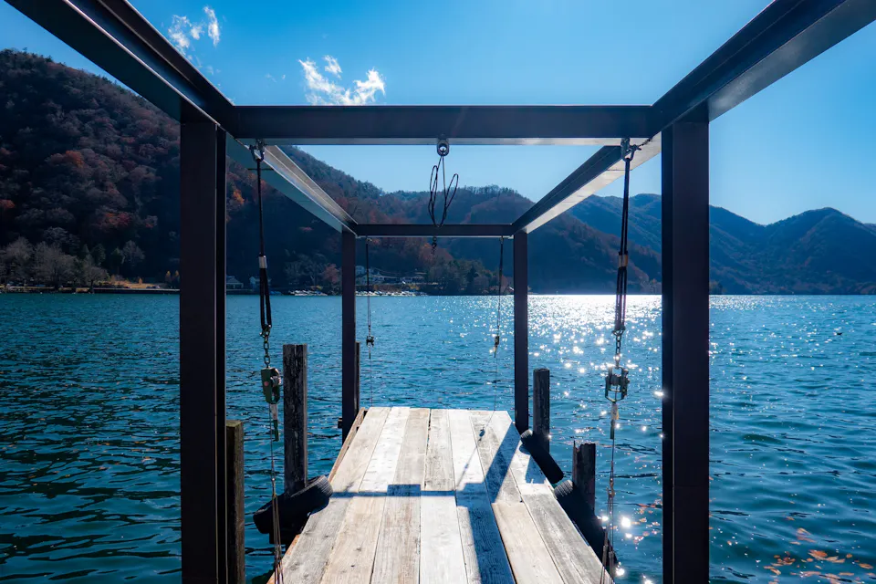 A wooden platform extends over a calm, sparkling lake, framed by a metal structure. Mountains under a clear blue sky provide a scenic backdrop. The sun's reflection shimmers on the water's surface, adding to the serene atmosphere.