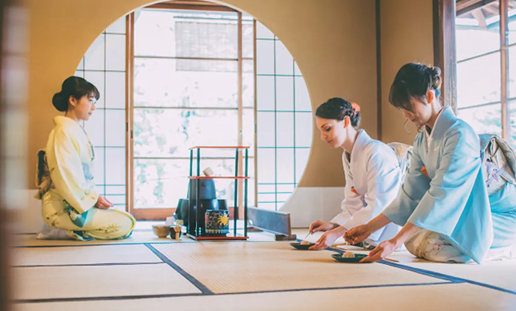 Tea Ceremony Three women in traditional Japanese kimonos participate in a tea ceremony in a room with shoji screen windows and tatami mats. Two women prepare tea on the floor, while one woman sits attentively, observing the process. Traditional utensils and decor are present.
