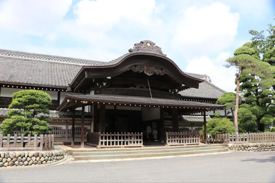 A traditional Japanese building with ornate wooden architecture and a curved roof stands under a partly cloudy sky. The entrance features a wooden canopy and a stone pathway, with neatly trimmed trees and a picket fence in the foreground.