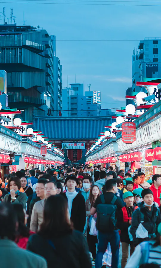 Nakamise Shopping Street A bustling outdoor market in Japan with crowds of people walking between rows of brightly lit shops and red lanterns, set against a backdrop of city buildings at dusk.