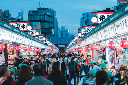 A bustling outdoor market in Japan with crowds of people walking between rows of brightly lit shops and red lanterns, set against a backdrop of city buildings at dusk.