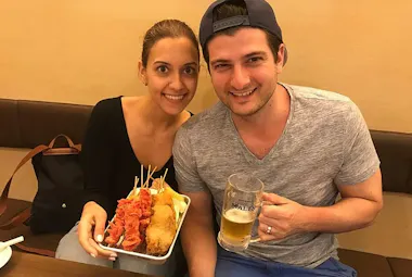 A smiling woman holds a tray of assorted skewered fried foods, while a smiling man beside her holds a glass mug of beer. They are seated together at a table in a casual indoor setting.