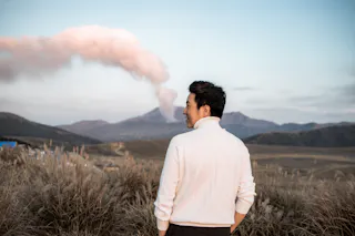 A man in a white sweater stands in a grassy field, looking over his shoulder toward a smoking volcano in the distance under a pale blue sky.