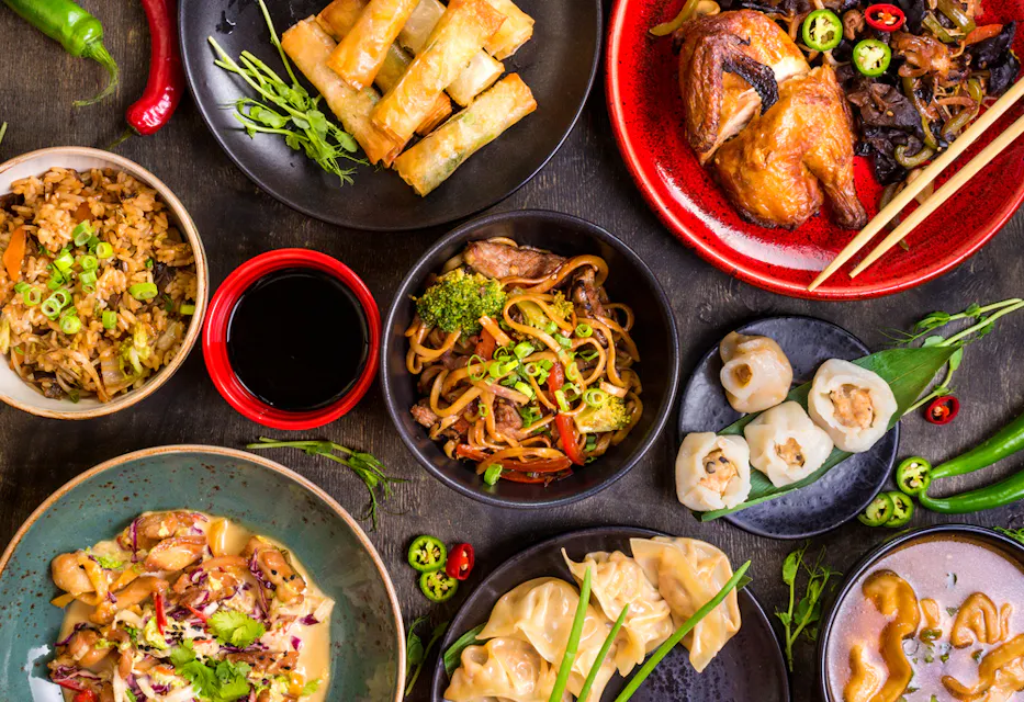 An assortment of Asian dishes including fried rice, spring rolls, dumplings, noodles with vegetables, chicken, and dipping sauces, presented in colorful plates and bowls on a dark wooden table, garnished with fresh herbs, chili slices, and green onions.