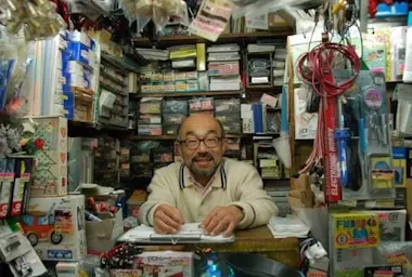 A smiling man with glasses stands behind a crowded counter in a small shop filled with various goods, boxes, and colorful items stacked on shelves and hanging from the ceiling.