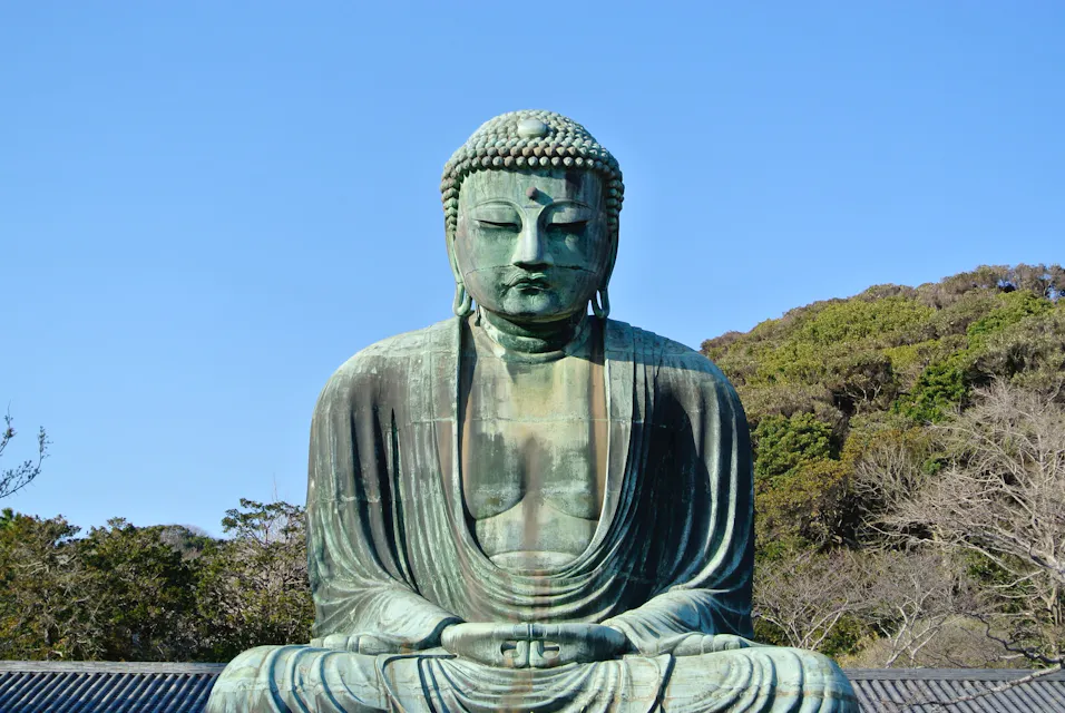 The image shows the Great Buddha of Kamakura, a large outdoor bronze statue of Amida Buddha located in Kamakura, Japan. The statue sits in a serene, meditative posture with a clear blue sky and lush greenery in the background.