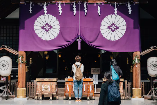 Three people stand in front of a traditional Japanese shrine, facing a large purple curtain adorned with white chrysanthemum emblems. The scene features wooden offertory boxes and traditional decorations.