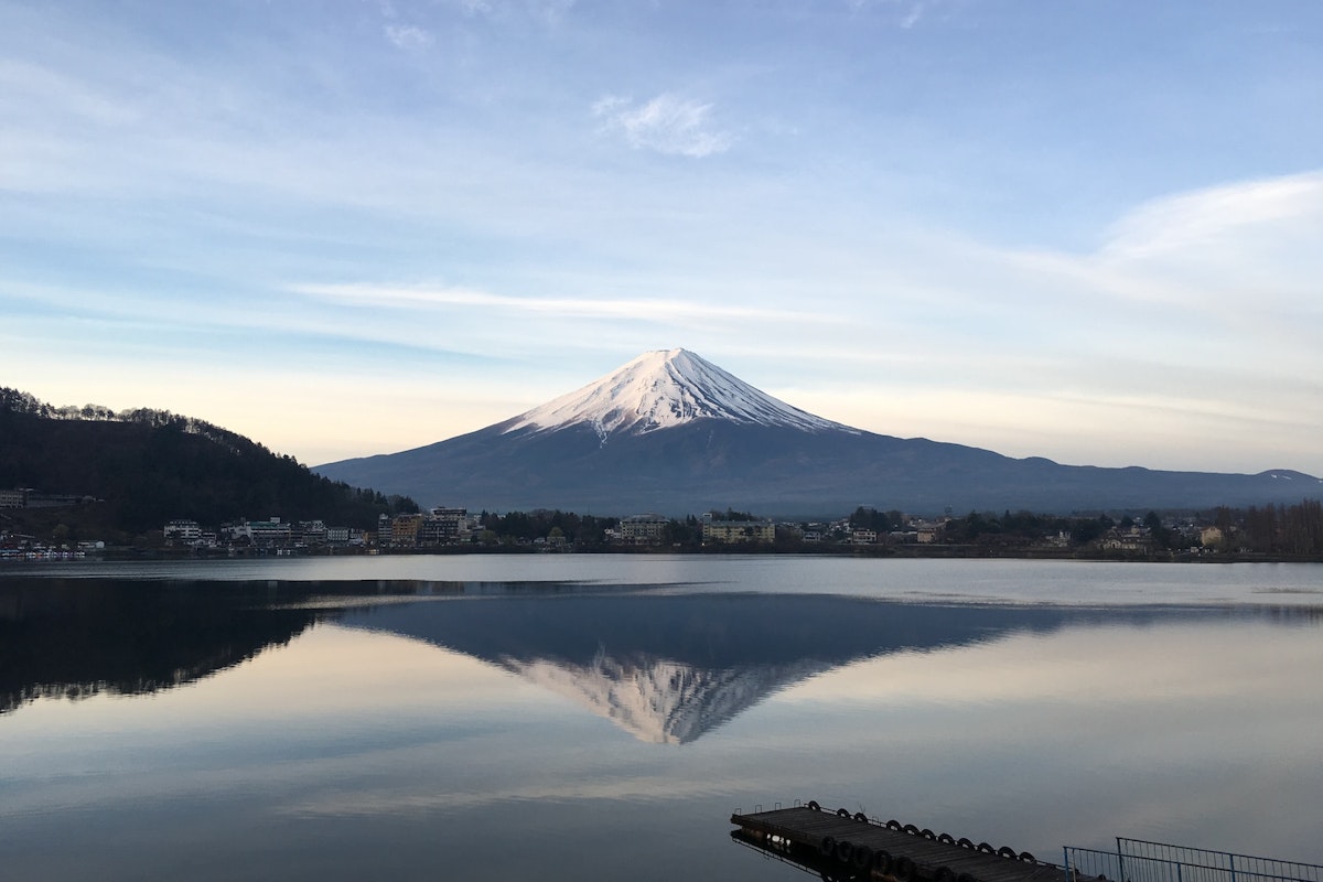 Mount Fuji A picturesque view of Mount Fuji with a snow-capped peak, reflected in the calm waters of a lake. The landscape includes surrounding hills and a few buildings at the base, all under a clear blue sky with subtle cloud formations.