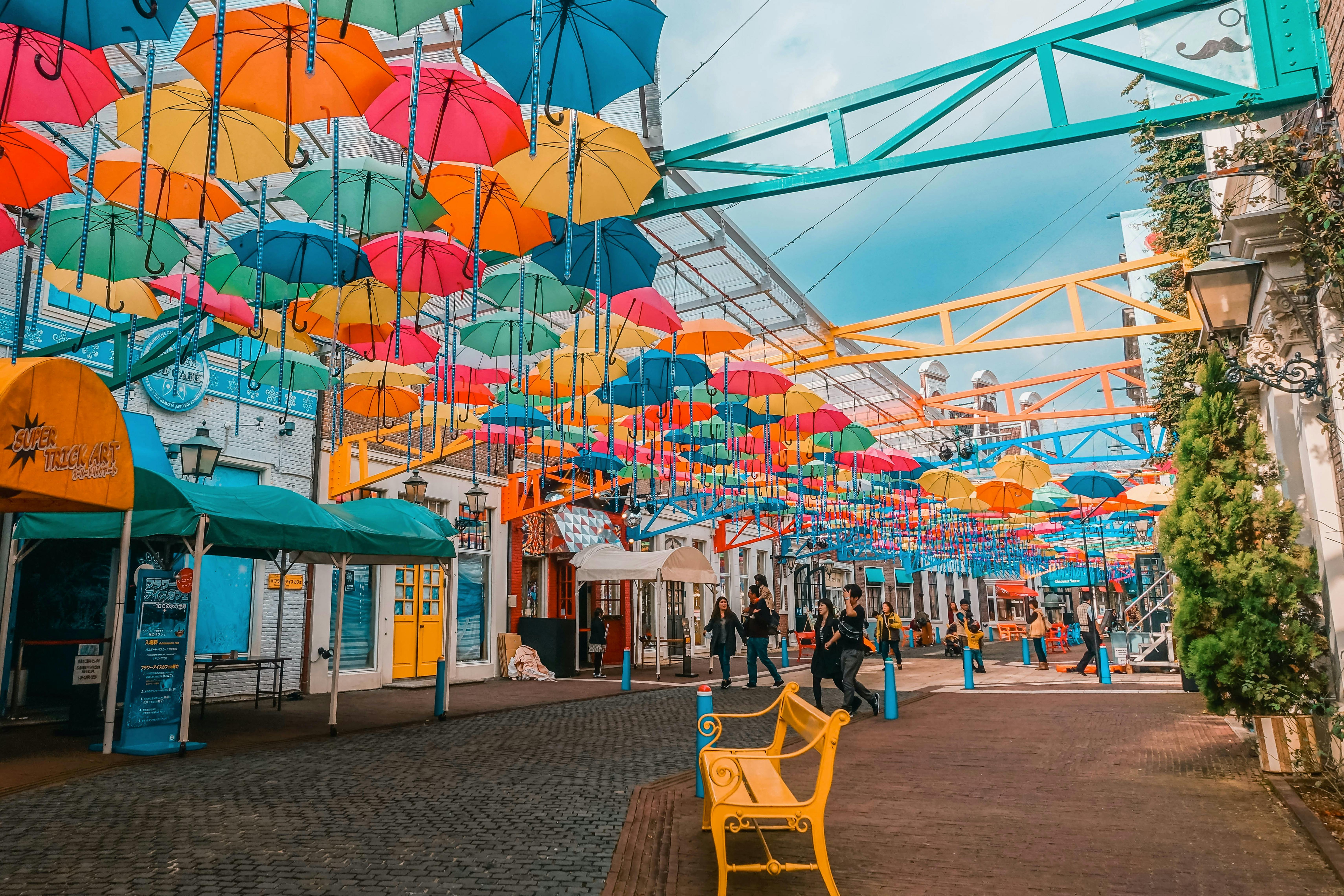 A colorful pedestrian street decorated with many hanging umbrellas in various bright colors. Yellow benches line the cobblestone path, and people walk under the cheerful canopy on a sunny day.