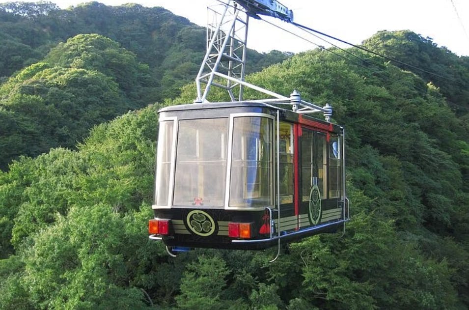 A cable car with large windows travels above a lush, green forested area, suspended by a cable. The cable car is decorated with traditional symbols and has metal frames on the roof.