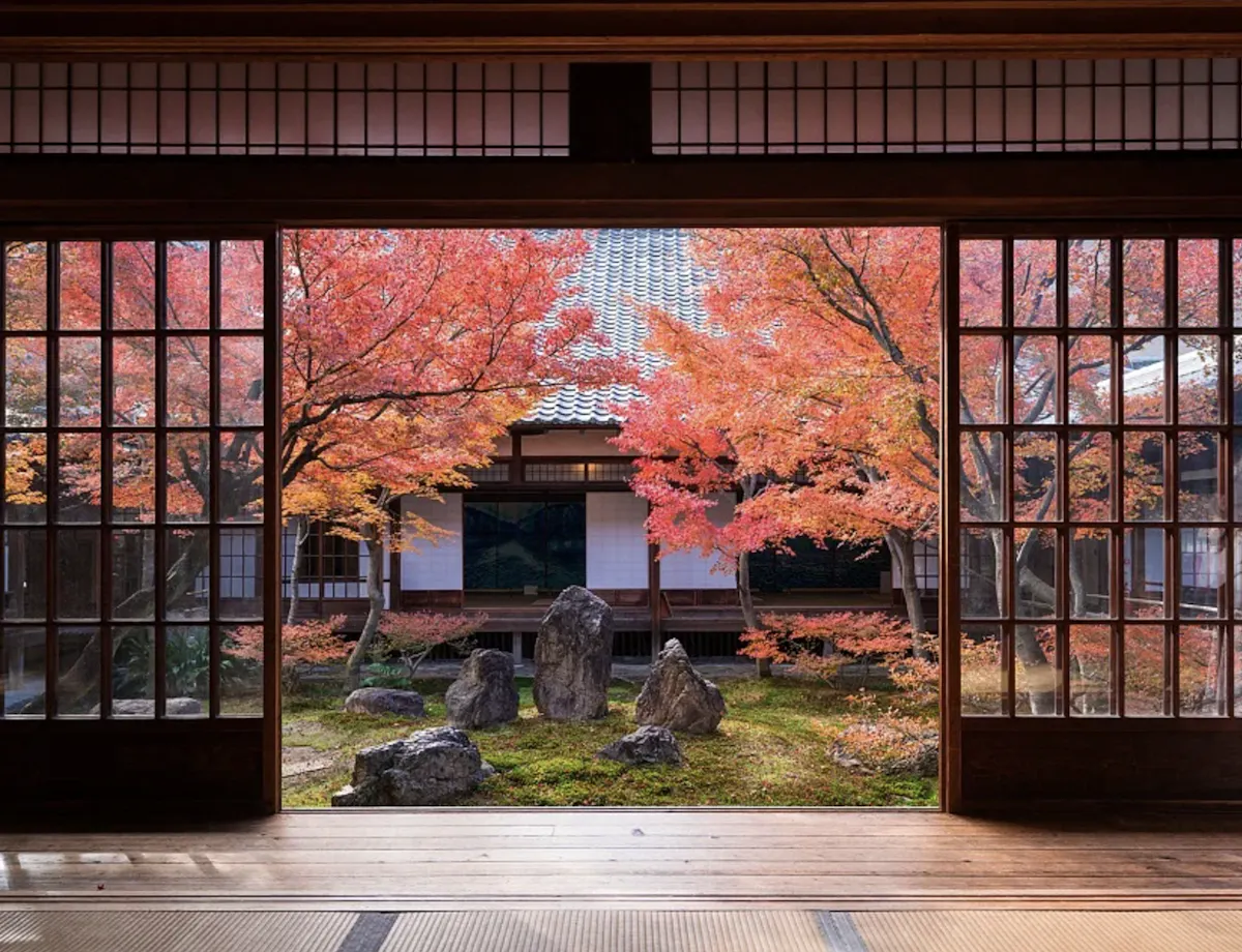 Gion Kyoto A traditional Japanese room with sliding wooden doors opens to a tranquil garden featuring autumn-colored maple trees and arranged rocks, with a building in the background.