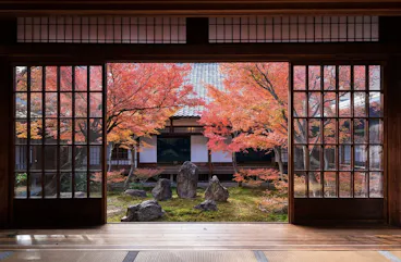 A traditional Japanese room with sliding wooden doors opens to a tranquil garden featuring autumn-colored maple trees and arranged rocks, with a building in the background.