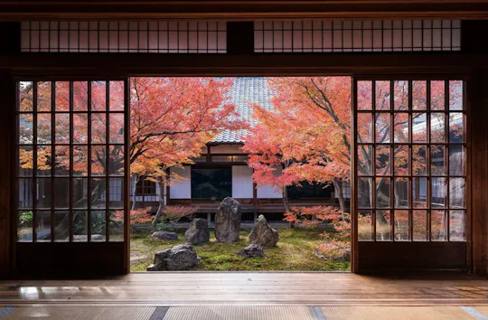 A traditional Japanese room with sliding wooden doors opens to a tranquil garden featuring autumn-colored maple trees and arranged rocks, with a building in the background.