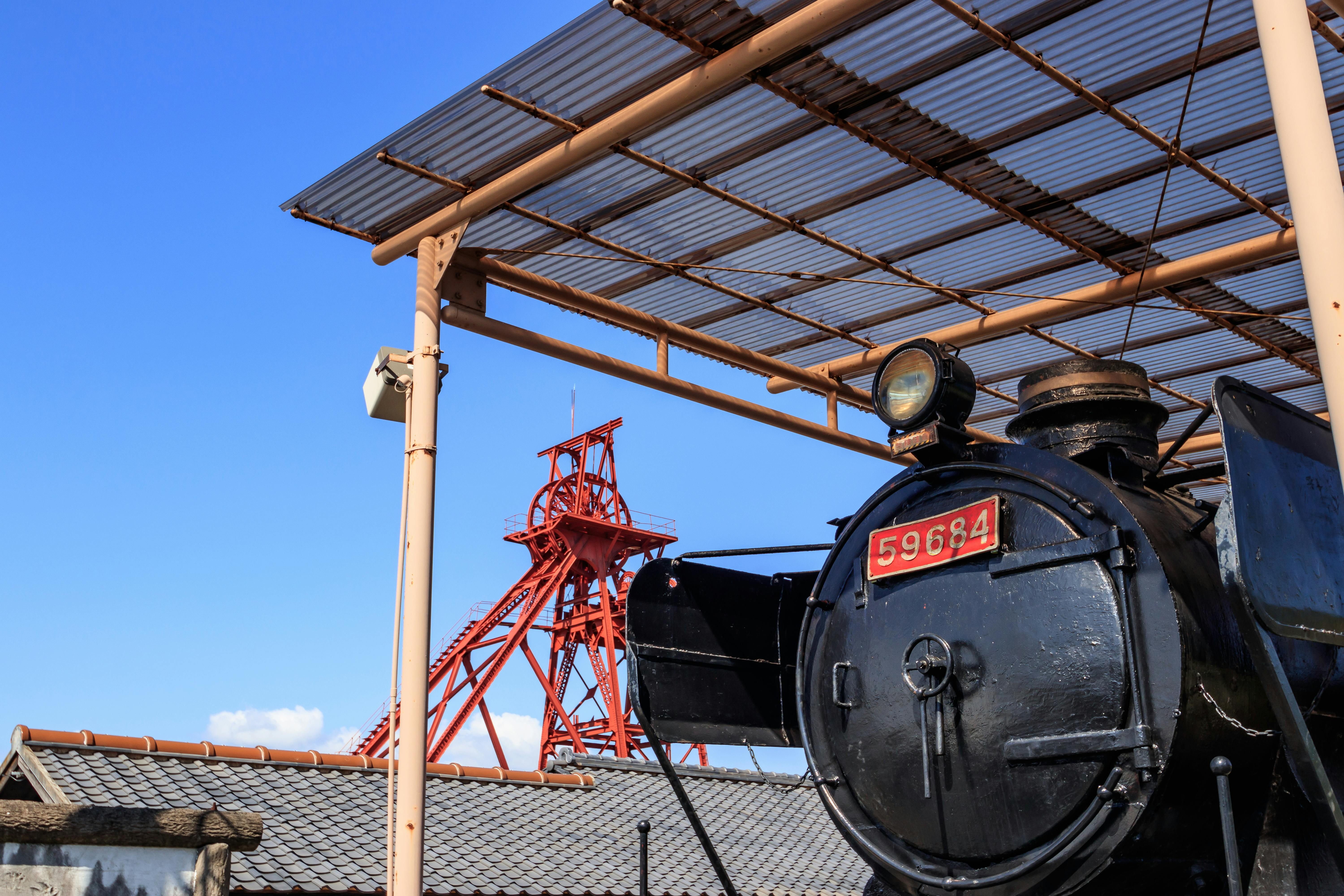 A black steam locomotive with the number 59684 under a metal canopy, with a red industrial steel structure and tiled rooftops in the background against a clear blue sky.