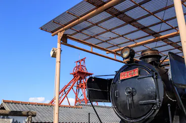 A vintage black steam locomotive with the number 59684 is displayed under a metal roof. In the background, a tall red metal mining structure rises against a clear blue sky.