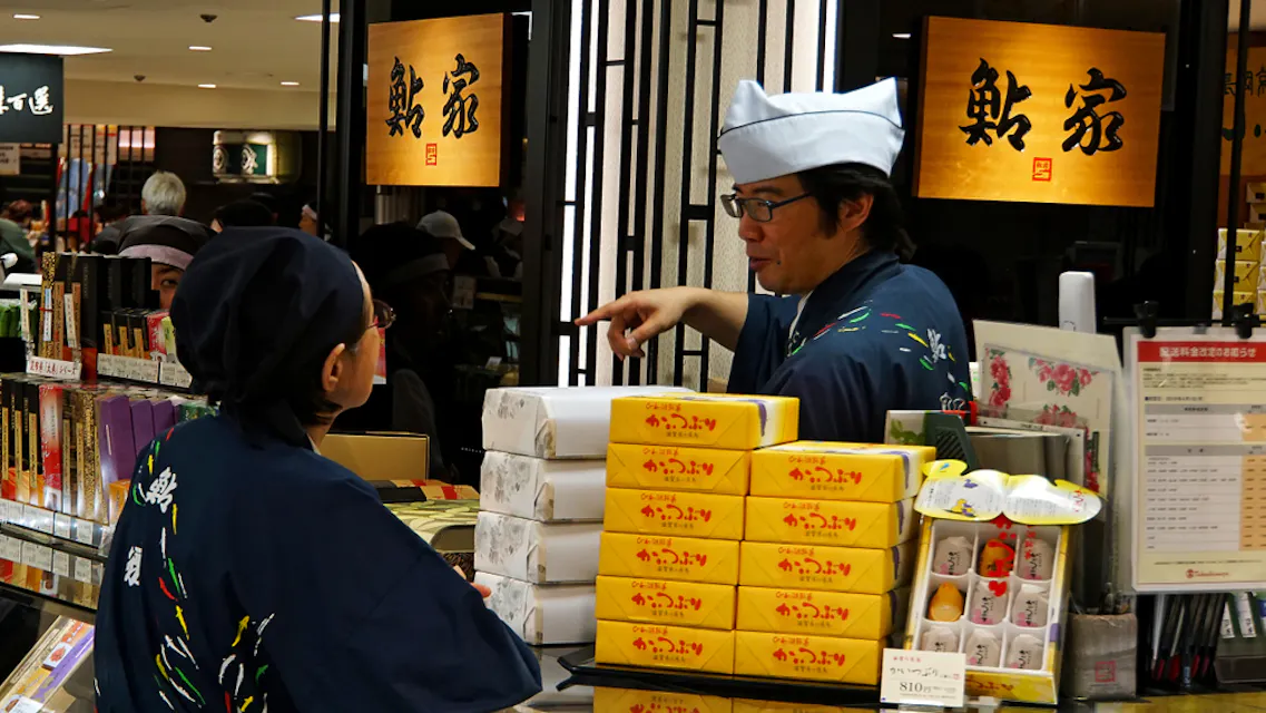 Two store employees, dressed in traditional Japanese attire with headgear, are seen talking behind a counter stacked with yellow boxes and various products. Japanese signs hang above them. The setting appears to be a busy market or store.
