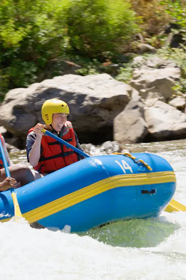 A person wearing a yellow helmet and red life jacket smiles while paddling in a blue inflatable raft on a river, with rocks and greenery in the background.
