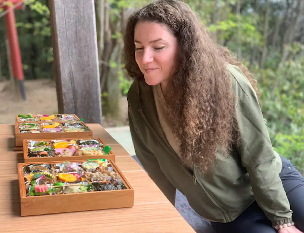 Eco-hiking in Hiroshima A woman with curly hair leans over a table outdoors, smiling at several colorful bento boxes filled with assorted Japanese food. Trees and greenery are visible in the background.