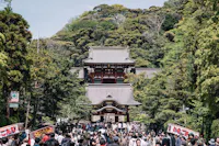 A bustling crowd walking towards Tsurugaoka Hachimangu Shrine in Kamakura, Japan. The main torii gate and pavilion of the shrine are visible amidst lush greenery. Colorful banners and signs flank both sides of the pathway, under a clear sky.