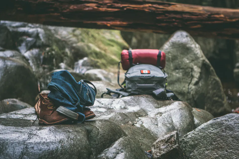 A pair of brown hiking boots, a rolled-up pair of blue jeans, and a grey backpack with a red sleeping bag attached are placed on large rocks in a rugged outdoor setting. A large fallen log is visible in the background with moss-covered boulders around.
