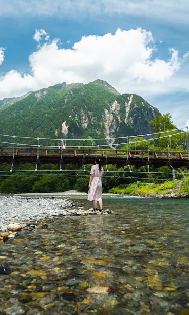 Kamikochi A person in a light dress stands on a rocky riverbank facing a wooden suspension bridge, with tall green mountains and a partly cloudy blue sky in the background.