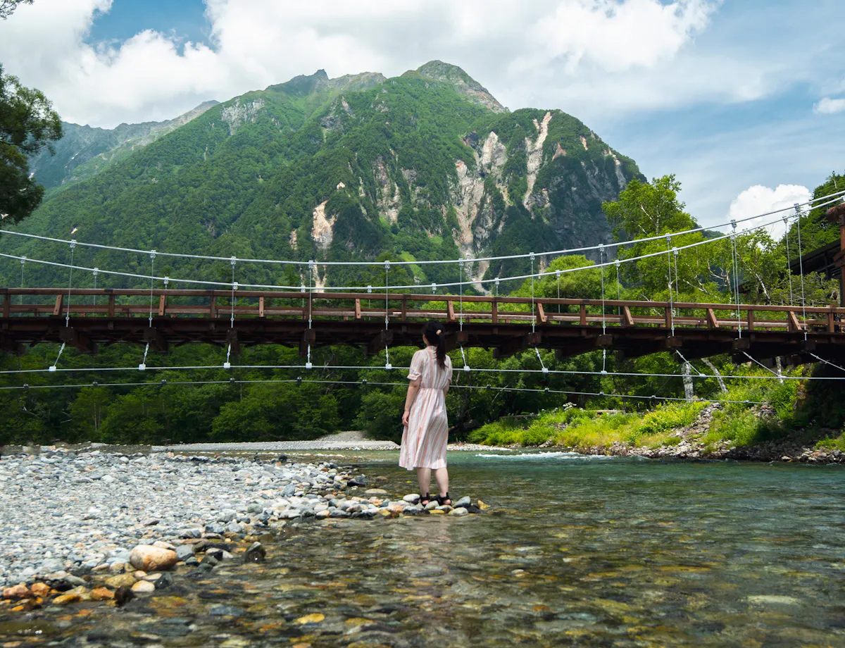 Kamikochi A person in a light dress stands on a rocky riverbank facing a wooden suspension bridge, with tall green mountains and a partly cloudy blue sky in the background.